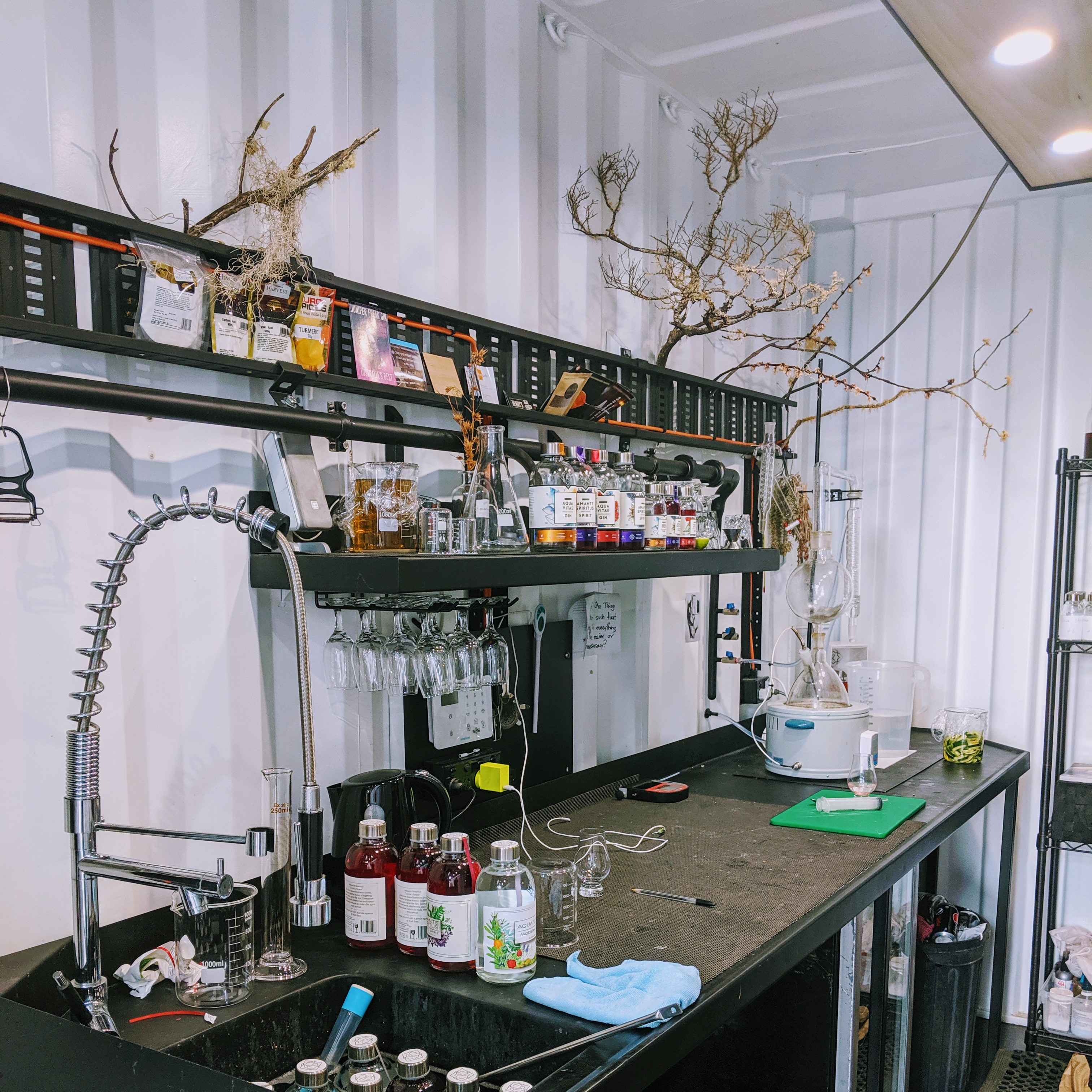 A laboratory style bench inside the shipping container that houses 7K Distillery. Clusters over brightly coloured Aqua Vitae gin bottles sit on the bench and shelving above. Dead tree branches covered in moss are artfully arranged to hang over the bench