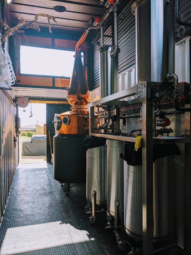 A copper still with black cladding sits next to a stainless steel gin column inside a shipping container